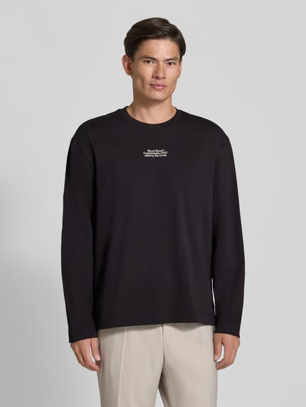 Mężczyzna nosi bluzkę z długim rękawem z bawełny, krój regular fit, Regular Fit Longsleeve aus reiner Baumwolle Modell 'Ryan' od Wood Wood, czarny kolor, wyhaftowane logo, długość tyłu w rozmiarze M: 79 cm, Ganzkörperansicht von vorne.