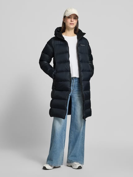 Frauenmodel trägt Steppmantel mit Daunen-Federn-Füllung Modell 'FROST' von Peak Performance in Schwarz, mit Kapuze und Reißverschluss, Ganzkörperansicht von vorne.