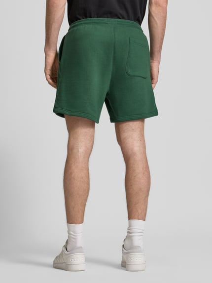 Männermodel trägt Regular Fit Shorts aus Baumwoll-Mix von Tommy Jeans in Grün, Optik: Unifarben, Sweat-Textur, Ganzkörperansicht Rückansicht.