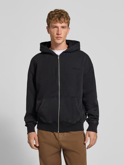 Männermodel trägt Regular Fit Sweatjacke aus Baumwoll-Mix von PEGADOR in Schwarz mit Kängurutasche und durchgehendem Zipper, Ganzkörperansicht von vorne. Kontext: Herren Sweatjacke, Logo-Stickerei, gerippte Abschlüsse, Rückenlänge 69 cm, Ärmellänge 67 cm.