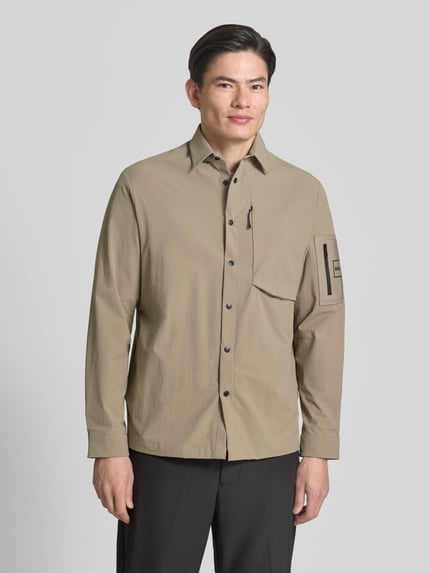 Męski model nosi Relaxed Fit Overshirt mit Label Patch Modell 'ST_GLOBE HEXA L' von BOSS Green, efekt: jednolity kolor, krój relaxed fit, wykładany kołnierz, listwa z zatrzaskami, aplikacja z metką, długi rękaw, hydrofobowy, wpuszczane kieszenie, brustasza, ganzkörperansicht von vorne.