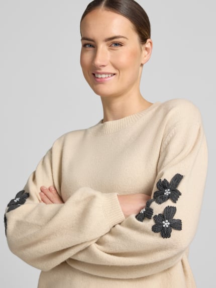 Frauenmodel trägt Strickpullover aus Wolle mit Stitchings und Perlen von Christian Berg Woman, beiger Farbton, schwarze Blumenapplikationen, Relaxed Fit, Nahaufnahme Oberkörper.