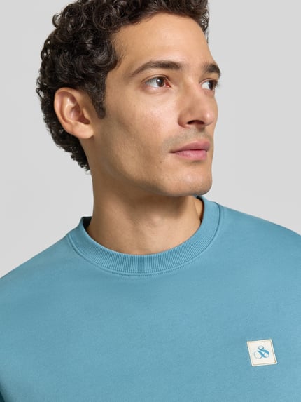 Männermodel trägt Sweatshirt mit Label-Patch von Scotch & Soda in hellem Blau, reine Baumwolle, lockerer Schnitt, Nahaufnahme Oberkörper.