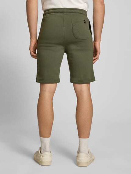 Männermodel trägt Shorts mit Eingrifftaschen und Logo-Print von Alpha Industries in Olivgrün, entspannter Schnitt, Rückansicht Ganzkörperansicht. Baumwoll-Mix, elastischer Bund mit Kordelzug, Gesäßtasche, glatte Textur, Größe M mit 39 cm Bundweite und 24 cm Innenbeinlänge.