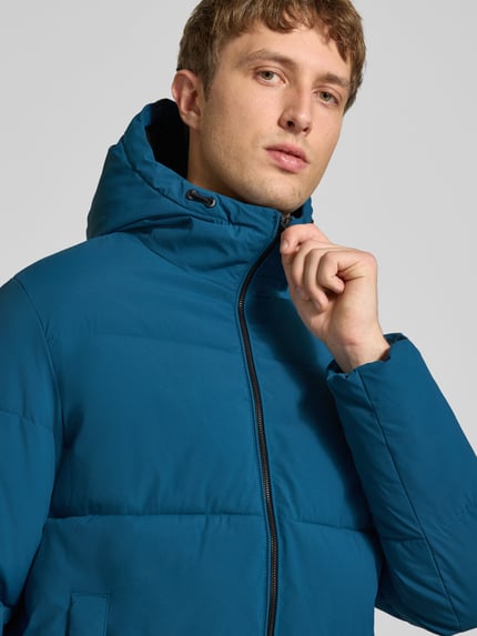 Męski model nosi kurtkę pikowaną z kapturem Model 'GALAXY' od JACK & JONES, kolor niebieski, zamek błyskawiczny, wpuszczane kieszenie, krój regular fit, detal: długość tyłu 70 cm, szerokość ramion 49 cm, szerokość w rozmiarze M 58 cm, długość rękawów 69 cm, zbliżenie na górną część ciała.