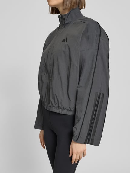 Frauenmodel trägt Regular Fit Jacke mit Stehkragen von adidas Sportswear, grau mit Logo-Print, Reißverschluss, elastische Abschlüsse, Nahaufnahme Oberkörper.