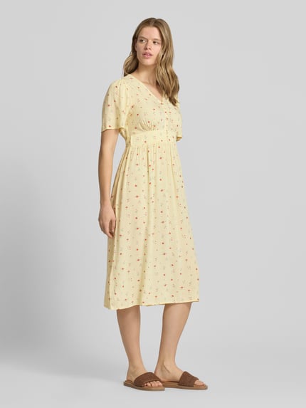 Vrouwenmodel draagt A-lijn midijurk van pure viscose, model 'TALA' van Pieces met all-over print, elastiek, korte knoopsluiting, V-hals, korte mouw, recht zoom; zachte gele kleur met kleine bloemmotieven; enkelvoudige midi lengte, luchtig materiaal; volledige lichaamsaanzicht van voren.