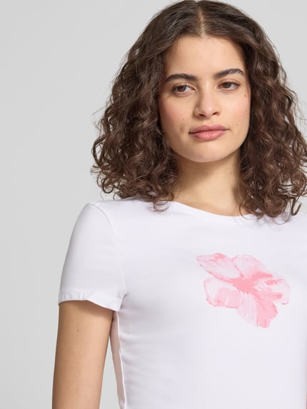 Vrouwenmodel draagt schmal geschnittenes T-Shirt aus Baumwollmix mit Blumen-Print van REVIEW in wit met roze bloem, nauwsluitende fit, ronde hals, korte mouwen, ruglengte maat S 54 cm, close-up bovenlichaam.
