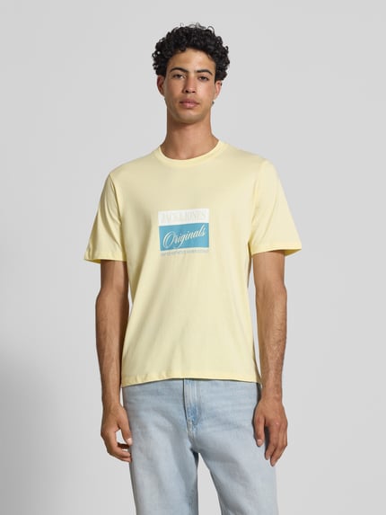 Męski model nosi t-shirt z nadrukiem z napisem model 'RHYERES' od Jack & Jones, czysta woskowana bawełna, krój regular fit, okrągły dekolt, długość tyłu w rozmiarze M: 67 cm, jasny żółty z niebiesko-białym nadrukiem, gładka tekstura, widok całej sylwetki z przodu.