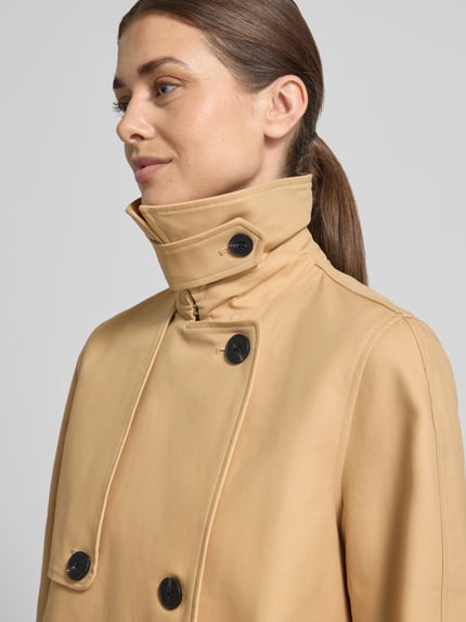Frauenmodel trägt Trenchcoat mit Ärmelriegeln von Stefanel in Beige mit schwarzen Knöpfen, reine Baumwolle, Nahaufnahme Oberkörper.