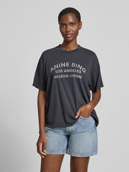 Kobiece model prezentuje Relaxed Fit T-Shirt mit Logo-Print od Anine Bing z czarnej bawełny z nadrukiem z logo, okrągły dekolt, krótki rękaw, długość tyłu w rozmiarze S: 59 cm. Ganzkörperansicht von vorne.
