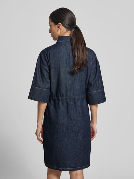 Frauenmodel trägt Knielanges Jeanskleid mit Brusttaschen von Comma aus Reine Baumwolle, Gerader-Schnitt, Knielänge, Hemdblusenkragen, Brusttasche, Knopfleiste, Bindegürtel in dunklem Jeansblau, rechteckige Form mit sichtbaren Nähten, glatte Textur, Ganzkörperansicht Rückansicht.