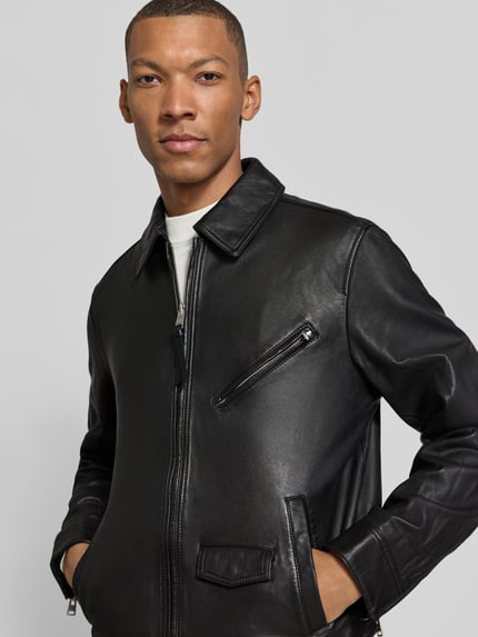 Męski model nosi Regular Fit Lederjacke aus Schafsleder od AllSaints, czarna skóra owcza, kołnierzyk typu kent, zamek błyskawiczny dwustronny, kieszeń na piersi zapinana na zamek, boczne i patkowe kieszenie, Model 'arto', półzbliżenie od przodu