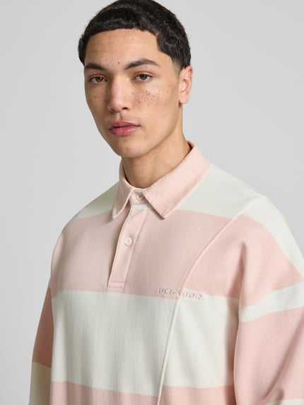 Männermodel trägt Relaxed Fit poloshirt uit puur katoen model 'Castelluccio' van Pegador met labelstitching, knoopsluiting, polokraag, mouwboordjes en overgeslagen schouders, kleur lichtroze-wit gestreept, context herenmode, ruglengte maat M 69 cm, detailopname bovenlichaam.