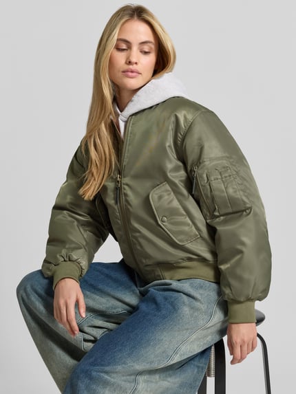 Frauenmodel trägt Bomberjacke mit Ärmeltasche Modell 'LEON' von Anine Bing in glänzendem Olivgrün, unifarben, gerippte Abschlüsse, Webware, Pattentaschen, Reißverschlusstasche, Ganzkörperansicht von vorne.
