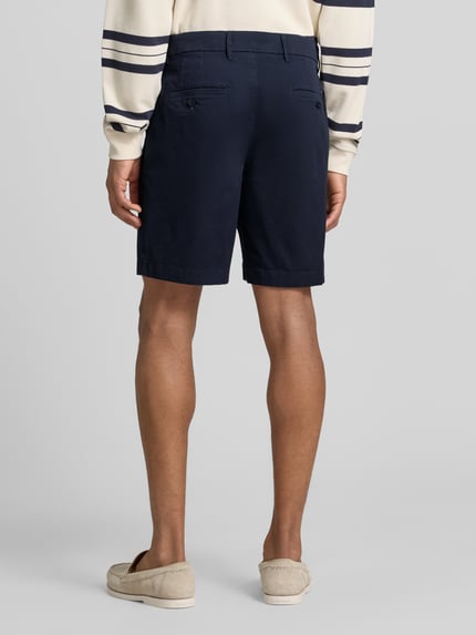 Männermodel trägt Chinoshorts aus Baumwoll-Mix Modell 'DOVER' von Tommy Hilfiger, Optik: Unifarben, Passform: Regular-Fit, Zwei Paspeltaschen mit Knopf hinten, Farbe Dunkelblau, feine glatte Textur, Ganzkörperansicht Rückansicht.