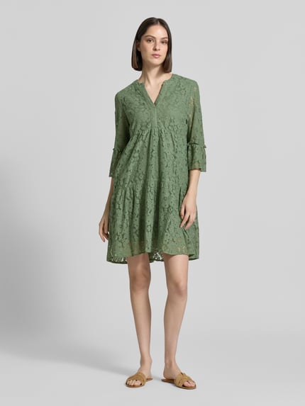 Frauenmodel trägt Loose Fit Spitzenkleid aus Baumwoll-Mix Modell 'HONEY' von Vero Moda in Olivgrün, Spitzenmuster, V-Ausschnitt, Glockenärmel, knielang, Ganzkörperansicht von vorne.