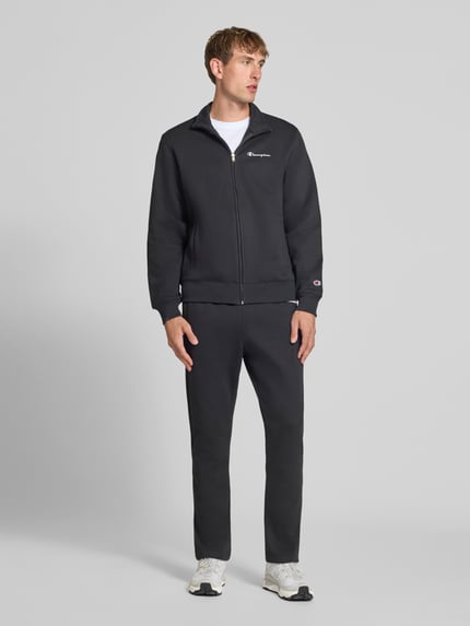 Męski model nosi Sweatanzug mit Label-Details od Champion, kolor czarny, mieszanka bawełny, prosty krój, stójka, zamek błyskawiczny, detal z logo, boczne wpuszczane kieszenie, elastyczny pas, Szerokość w rozmiarze M: 76,42 cm, Długość tyłu w rozmiarze M: 70 cm, Szerokość ramion w rozmiarze M: 51 cm, Długość rękawów w rozmiarze M: 69 cm, Długość wewnętrzna nogawki w rozmiarze M: 76 cm, widok całej sylwetki z przodu.