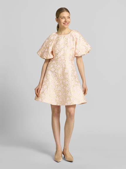 Frauenmodel trägt Tailliertes Cocktailkleid mit Allover-Muster Modell 'NORA' von Vero Moda, Farben in zartem Rosa, Mini-Länge, Rundhals-Ausschnitt, Schleifen-Verschluss, Puffärmel, Rückenausschnitt V-Ausschnitt, Optik:Allover-Muster, Passform: tailliert, Ganzkörperansicht von vorne.
