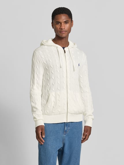 Männermodel trägt Regular Fit Strickjacke aus reiner Baumwolle von Polo Ralph Lauren in Weiß mit Zopfmuster, Ganzkörperansicht von vorne.