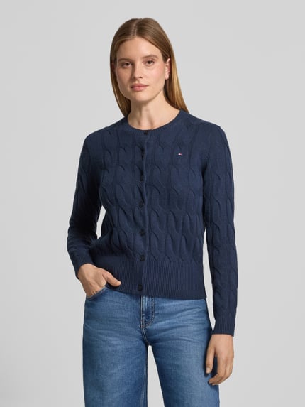 Frauenmodel trägt Strickjacke aus Wolle mit Logo-Detail von Tommy Hilfiger in Dunkelblau, Zopfmuster, Knopfleiste, Ganzkörperansicht von vorne.