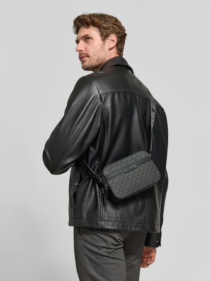 Mannenmodel draagt Camera Bag met Logo-applicatie Model 'Hudson' van Michael Kors in zwart structuurpatroon, hoofdvak met ritssluiting, verstelbare schouderband, breedte 22 cm, lengte 14 cm, diepte 6 cm, bandlengte 98 cm, kleurcontrast tussen tas en kleding, vooraanzicht, Nahaufnahme Oberkörper.