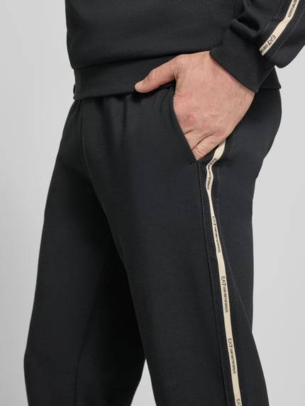 Männermodel trägt schwarze Sweatpants aus Baumwoll-Mix von EA7 Emporio Armani, beige Seitenstreifen, Eingrifftaschen, Nahaufnahme Oberkörper.