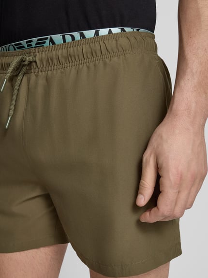 Männermodel trägt Relaxed Fit Badeshorts mit Logo-Stitching von Emporio Armani in Olive mit elastischem Bund und Tunnelzug, Nahaufnahme Oberkörper.
