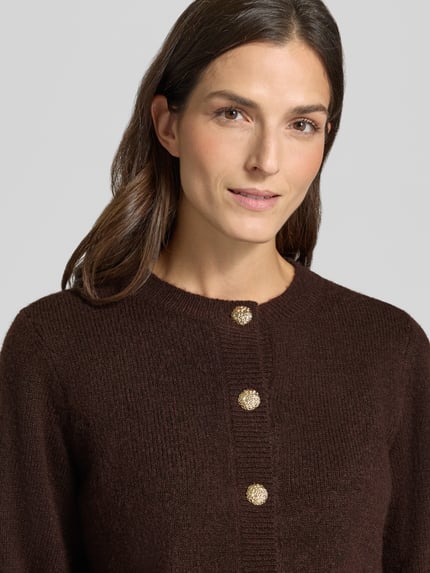 Frauenmodel trägt Strickjacke mit Zierknöpfen Modell 'VMHAZEL' von VERO MODA in Braun mit metallischen Akzenten, Nahaufnahme Oberkörper.