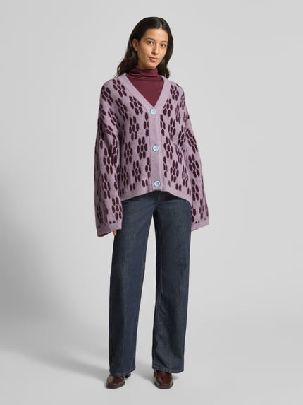 Frauenmodel trägt Strickjacke mit Knopfleiste Modell 'VITOW' von American Vintage im Alpaka-Lanawoll-Mix mit Stretch-Anteil, lockerer Schnitt, gerippte Abschlüsse, V-Ausschnitt, Muster in Lila- und Violett-Tönen, Rückenlänge ca. 67 cm, Ärmellänge ca. 53 cm, Ganzkörperansicht von vorne.