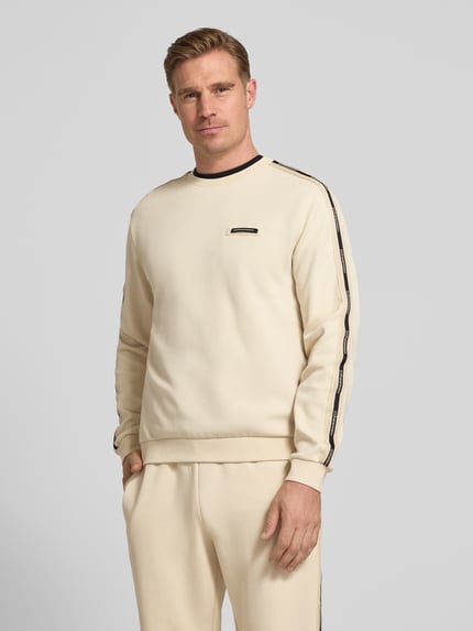Męski model nosi Sweatshirt mit Label-Details und Rundhalsausschnitt von EA7 Emporio Armani, mieszanka bawełny, krój regular fit, kremowy, detale z logo, okrągły dekolt, długość tyłu w rozmiarze M: 63 cm, długość rękawów w rozmiarze M: 66 cm, widok całej sylwetki z przodu.