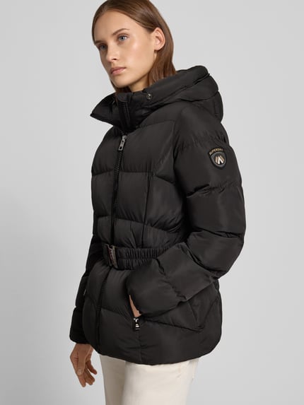 Frauenmodel trägt Mantel in Stepp-Optik Modell 'FUJI' von Superdry mit Kunstfellbesatz und Gürtel, taillierte Passform, Ganzkörperansicht von vorne.