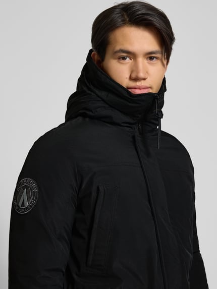 Männermodel trägt Parka mit Kapuze von Superdry in Schwarz, Material-Mix mit Stretch, Reißverschlusstaschen, Nahaufnahme Oberkörper.