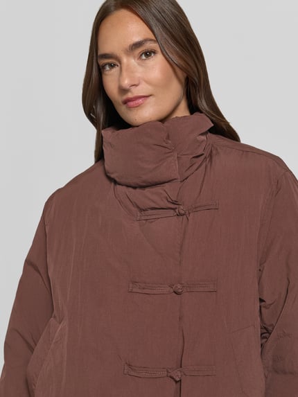 Frauenmodel trägt Jacke mit Stehkragen Modell 'TRUFA' von MANGO in Braun mit Reiß- und Schlaufenverschluss, Nahaufnahme Oberkörper. Gerader Schnitt, seitliche Eingrifftaschen, elastische Ärmelabschlüsse, sichtbare Textur, Kontext: Rückenlänge 61 cm, Schulterbreite 40 cm, Brustweite 52 cm, Ärmellänge 63 cm.