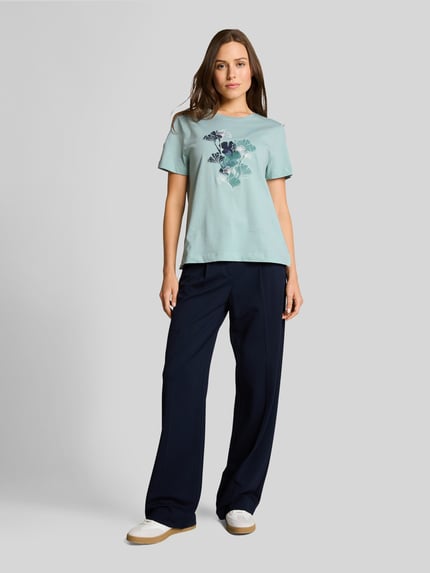 Frauenmodel trägt Regular Fit T-Shirt aus reiner Baumwolle von Tom Tailor in Mint mit floralem Motiv-Print und kurzem Arm, kombiniert zu dunkelblauer Hose, unifarbene Optik, Jerseytextur, Ganzkörperansicht von vorne.