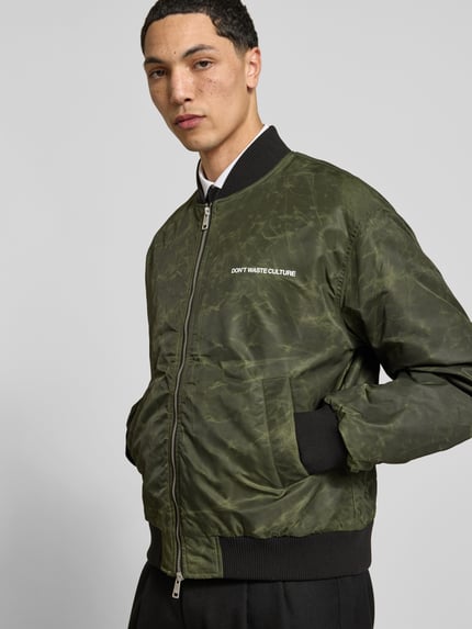 Męski model nosi bomberkę z nadrukiem z logo 'Bomberjacke mit Label-Print' od Don't Waste Culture w kolorze khaki, krój regular fit, zamek błyskawiczny dwustronny, elastyczne wykończenia, stójka, długość tyłu w rozmiarze M: 67 cm, szerokość ramion w rozmiarze M: 53 cm, długość rękawów w rozmiarze M: 64 cm, zdjęcie: całkowity widok z przodu.