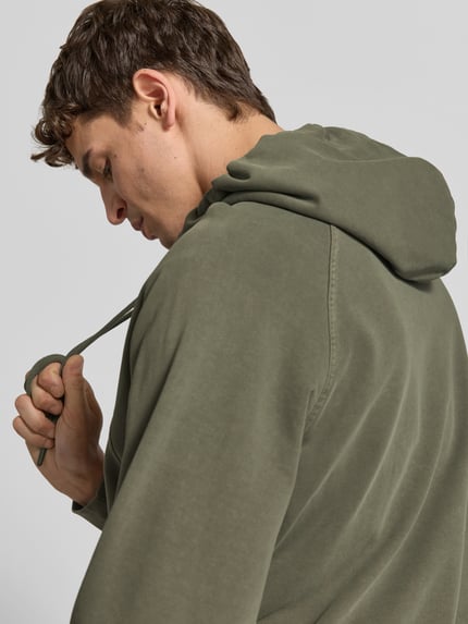 Männermodel trägt Regular Fit Hoodie aus reiner Baumwolle von Closed in Olivgrün, Kapuze mit Tunnelzug, Nahaufnahme Oberkörper, krój regular fit, 62 cm długość z tyłu w rozmiarze M, elastyczne wykończenia, kieszeń kangurka