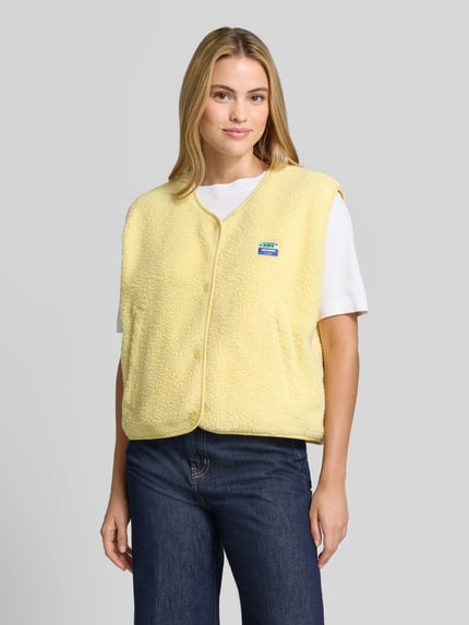 Frauenmodel trägt Relaxed Fit Weste aus Teddyfell Modell 'Hoktown' von American Vintage, gelb, Knopfleiste, Logo-Patch, Nahaufnahme Oberkörper. Damen Weste, Rückenlänge 56 cm, Schulterbreite 43 cm, Brustweite 63 cm.