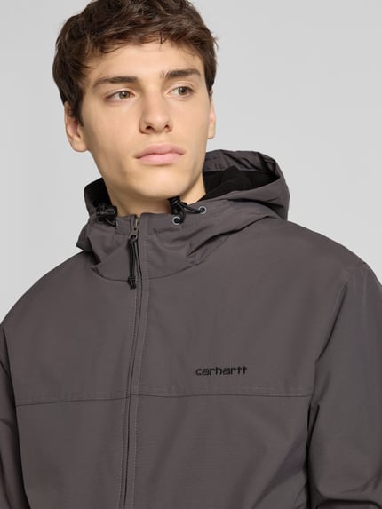 Männermodel trägt Jacke mit Kapuze von Carhartt Work In Progress in Grau mit Reißverschluss, Label-Stitching, Nahaufnahme Oberkörper.