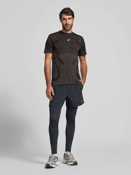 Męski model nosi męski t-shirt Slim Fit RUNNING T-Shirt Model 'ROAD' od ASICS, okrągły dekolt, nadruk z logo w efekcie metalicznym, długość tyłu w rozmiarze M: 72 cm, ciemne odcienie, gładka faktura, widok całej sylwetki z przodu.
