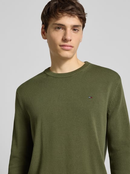 Männermodel trägt Regular Fit Strickpullover aus reiner Baumwolle von Tommy Jeans in Olivgrün, Rundhals, unifarben, Nahaufnahme Oberkörper.