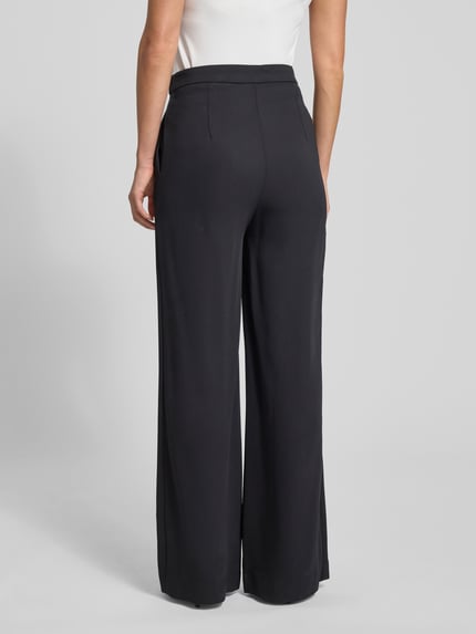 Männermodel trägt czarne, szerokie spodnie z wiskozy Wide Fit Stoffhose aus Viskose-Mix Modell 'Inga' od GUIDO MARIA KRETSCHMER WOMAN, fałdy, zapięcie na haftkę i zamek, materiał elastyczny, widok całej sylwetki od tyłu.