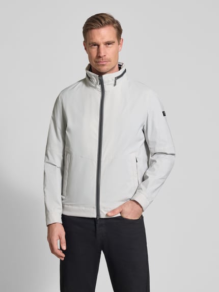 Männermodel trägt Regular Fit Jacke mit Stehkragen von bugatti in hellem Ton, mit Logo-Detail, Zweiwege-Reißverschluss, Eingrifftaschen, elastische Ärmelabschlüsse, verstaubarer Kapuze, Ganzkörperansicht von vorne. Rückenlänge 68 cm, Schulterbreite 42 cm, Brustweite 50 cm, Ärmellänge 65 cm.