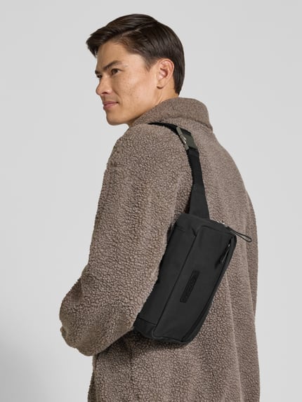Männermodel trägt schwarze Bauchtasche mit Crossbody-Tragegurt aus reiner Baumwolle von Marc O'Polo, unifarben, Canvas-Struktur, Ganzkörperansicht Rückansicht.