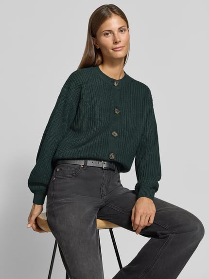 Modelka nosi zielony kardigan rozpinany Loose Fit Strickjacke mit einreihiger Knopfleiste model 'LEA' von VERO MODA, damska, faktura dzianiny, widok całego tułowia z przodu.