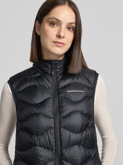 Frauenmodel trägt Steppweste mit Daunen-Federn-Füllung Modell 'HELIUM' von Peak Performance in Schwarz, horizontale Steppnähte, glänzende glatte Textur, Logo-Stitching, Nahaufnahme Oberkörper. Slim Fit, Stehkragen, Reißverschluss, Damen Steppweste Kontext integriert.
