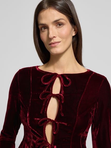 Frauenmodel trägt Bluse aus Samt mit Schürung Modell 'XARANJA' von Mango in dunklem Rot, taillierter Schnitt, Rundhalsausschnitt, Nahaufnahme Oberkörper. Damen Bluse, Rückenlänge 48 cm, Ärmellänge 62 cm, glänzende Samt-Textur.