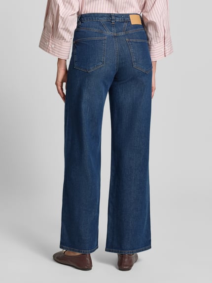 Modelka nosi Loose Fit Jeans aus Baumwoll-Mix Modell 'MADINA WIDE' od Tom Tailor o optyce denim, w kroju loose fit, z wysokim stanem, 5-kieszeniowym designem, naszywką z logo, zapięciem na guzik i kryty zamek, tkanina twill, widok całej sylwetki z tyłu.