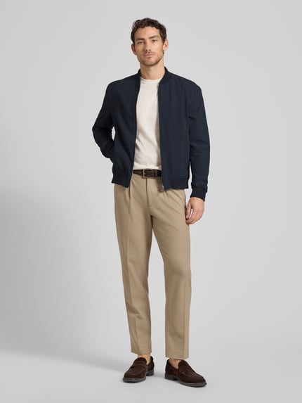 Mannelijk model draagt bomberjack met steekzakken model 'Indro' van JOOP! collectie, materiaalmix met stretch, recht model, opstaande kraag, ritssluiting, ribboorden, marineblauw, lichte broek, geheel aanzicht van voren.