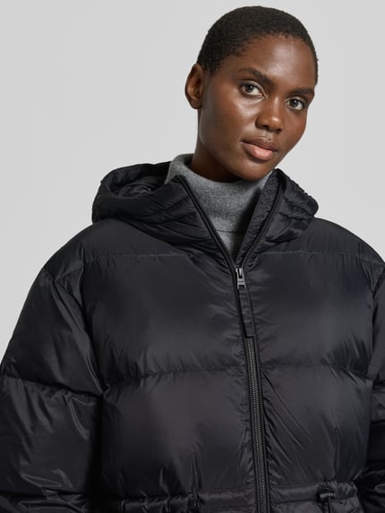 Kobieta modelka nosi czarną kurtkę 'Jacke mit Kapuze und Zweiwege-Reißverschlus' od Closed, srebrnoszary golf, tkanina błyszcząca, tekstura pikowana, prosty krój, kaptur, trok i zamek błyskawiczny dwustronny, wpuszczane kieszenie, dzianinowe zakończenia rękawów, szerokość w rozmiarze S: 62 cm, długość tyłu w rozmiarze S: 77 cm, szerokość ramion w rozmiarze S: 53 cm, długość rękawów w rozmiarze S: 63 cm, ujęcie: zbliżenie na górną część ciała.